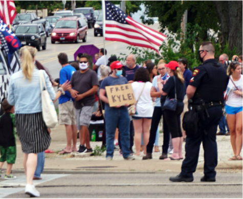A man is holding a sign that says Free Kyle on the streets. A policeman observes, and an American flag is visible
