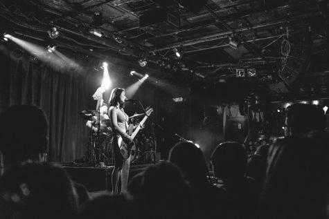 Mitski is singing while playing the guitar in front of a live audience in a rock club. The photo is in black and white.