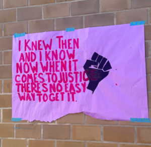 A purple-background poster is taped onto school walls. The poster says, I knew then, and I know now when it comes to justice, theres no last way to get it, in red letters. A symbol shows a black fist held up.
