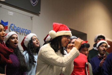 A girl caroling in a Santa hat mimes a trumpet.