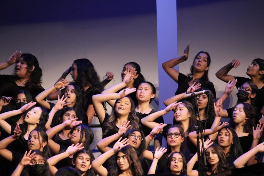 Choir students in black formal wear sing while standing on risers on a stage, placing the back of a palm to their foreheads, representing stress or weariness.