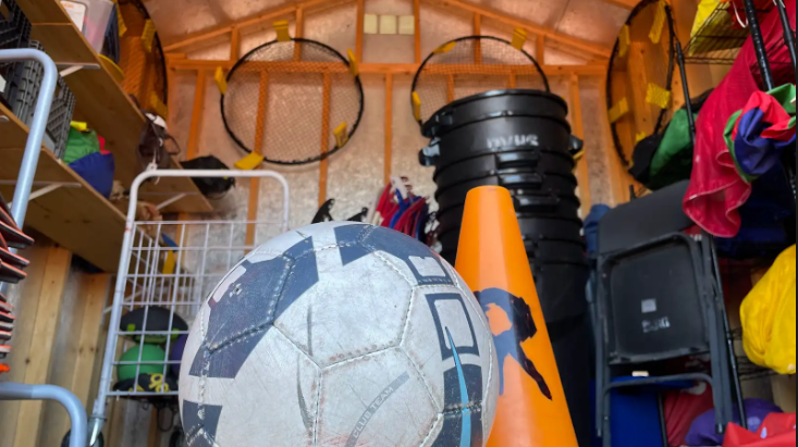 : A view of the PE shed from the ground, with a soccer ball and a plastic orange cone at the front. Equipment like this is managed by the PE program, and is paid for by the school budget. For sports, that’s not the case.